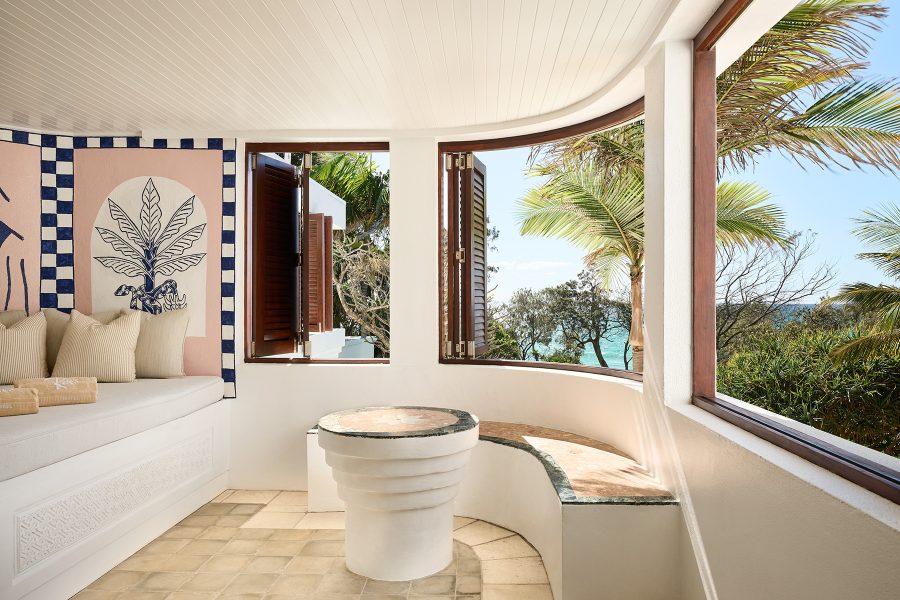 A curved white bathroom with travertine bench, looking out to palms and ocean.